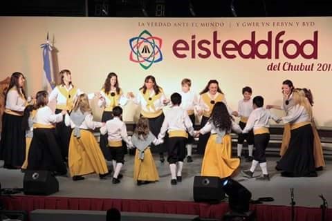Niños y jóvenes bailan en ronda frente al cartel del Eisteddfod